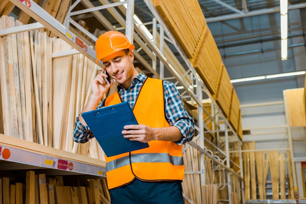 улыбающийся работник склада разговаривает на смартфоне и смотрит в буфер обмена
 - Фото, изображение