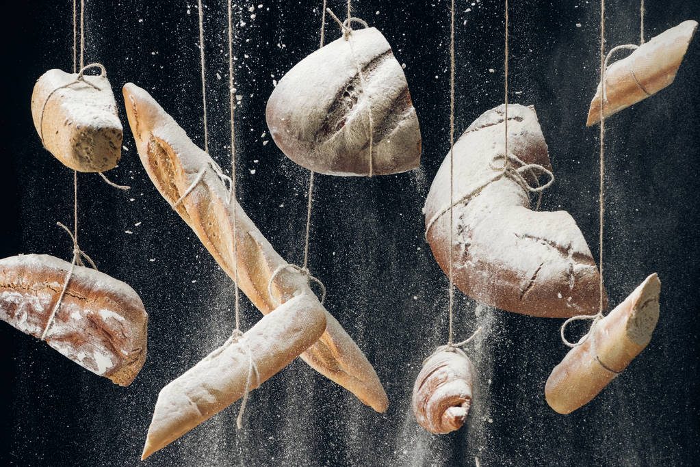 white flour falling at baked bread, baguettes and croissant hanging on ropes on black background - Photo, Image