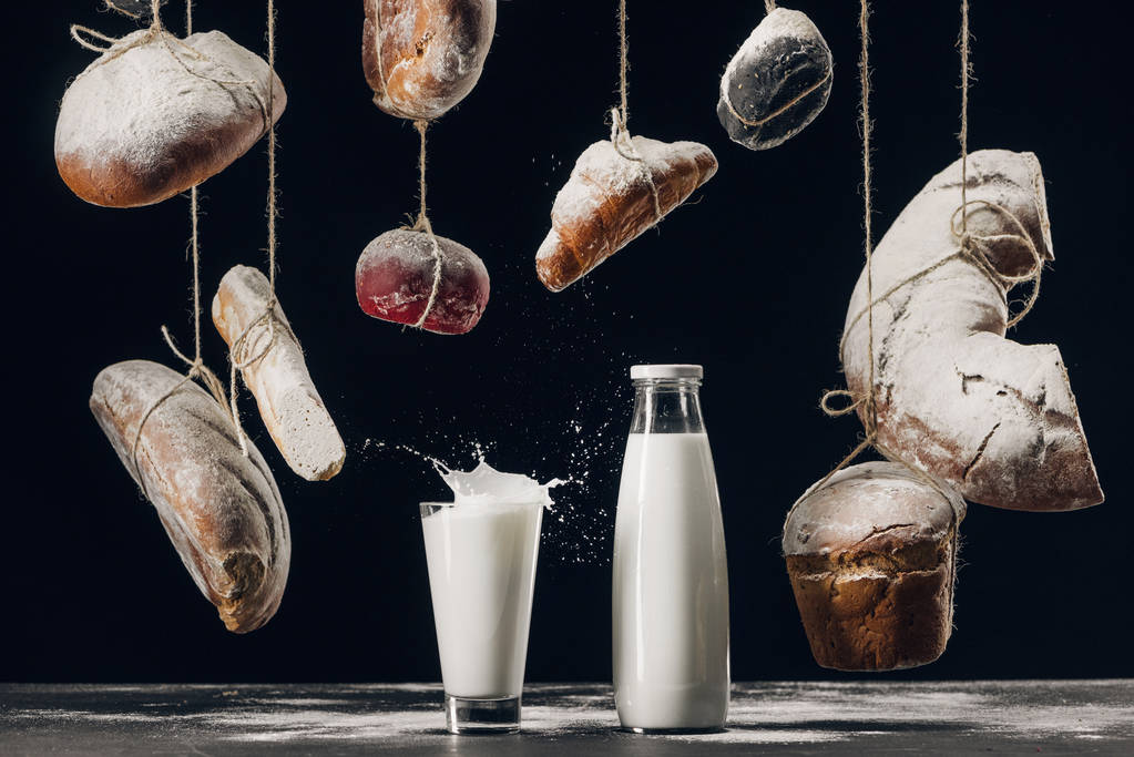 Milch spritzt im Glas auf den Tisch und Brot mit Mehl hängt an Schnüren isoliert auf schwarz - Foto, Bild