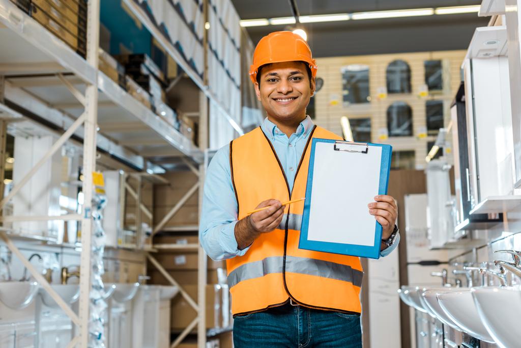 smiling indian warehouse worker showing clipboard with blank paper and looking at camera - Photo, Image