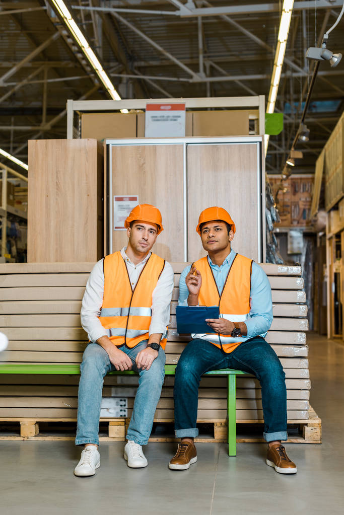 přemýšliví multikulturní pracovníci v bezpečnostních vesticích a přilbách sedícího na lavičce ve skladu  - Fotografie, Obrázek