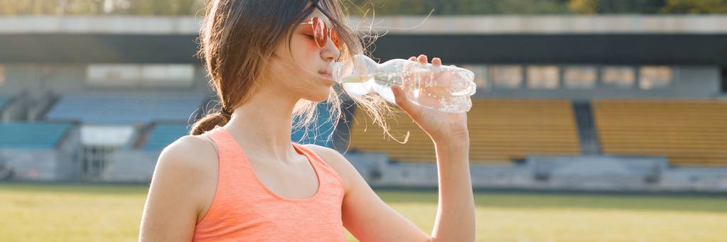 Jovencita adolescente bebiendo agua de la botella después de correr en el estadio, pancarta panorámica en la soleada noche de primavera
 - Foto, Imagen