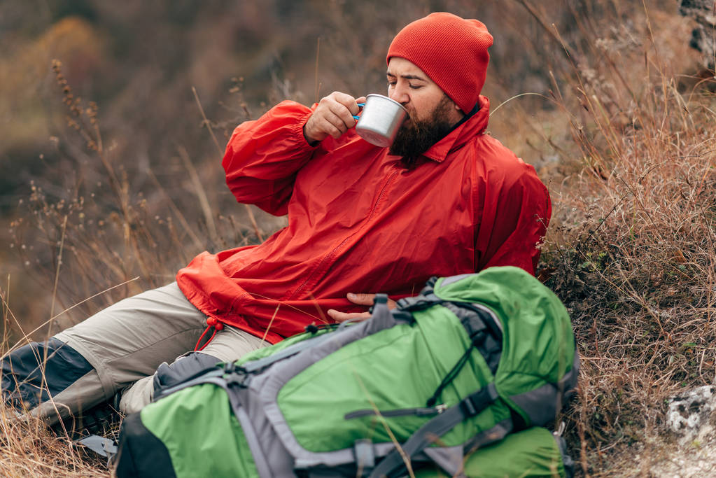 junger Entdecker Mann trinkt Heißgetränk in den Bergen, sitzt und entspannt sich nach dem Trekking. Reisender Mann mit rotem Hut, der nach dem Wandern eine Tasse Tee in der Hand hält. Reisen, Menschen - Foto, Bild