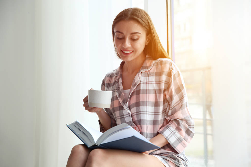 Jeune femme avec tasse de café livre de lecture près de la fenêtre à la maison
 - Photo, image