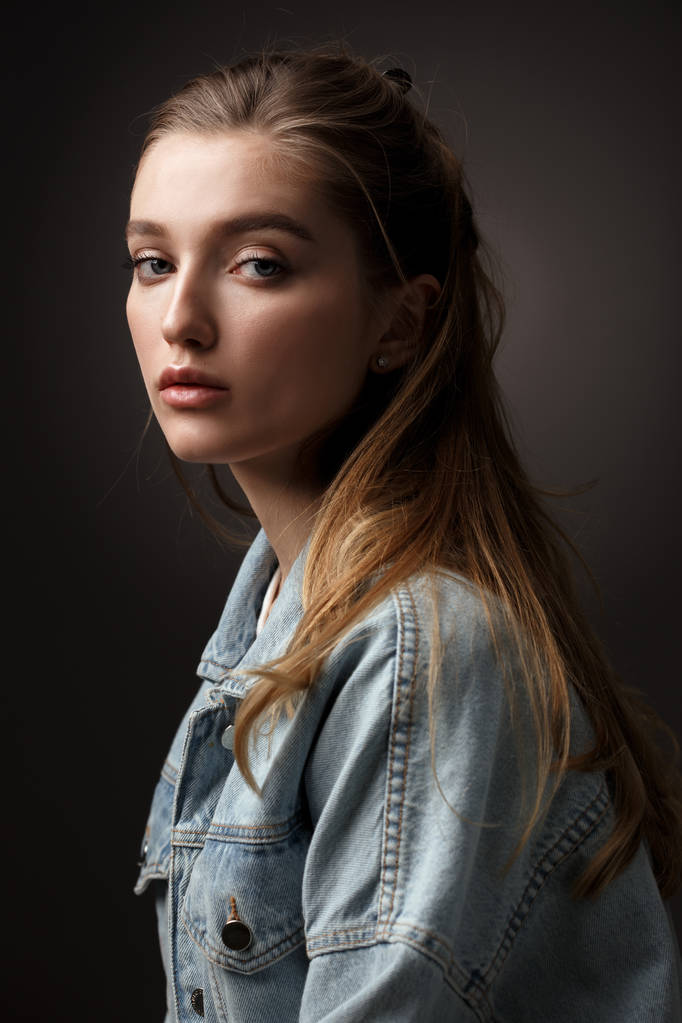 Retrato de bela menina morena com cabelo amarrado de volta vestida com jaqueta de jeans no fundo escuro no estúdio
 - Foto, Imagem