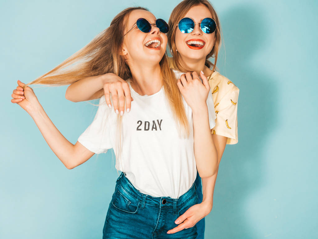 Dwie młode piękne, uśmiechnięte dziewczyny blond hipster w modne letnie kolorowe koszulki odzież. Sexy kobiety beztroski, pozować w pobliżu niebieską ścianą w okulary przeciwsłoneczne. Pozytywnych wzorców zabawy - Zdjęcie, obraz