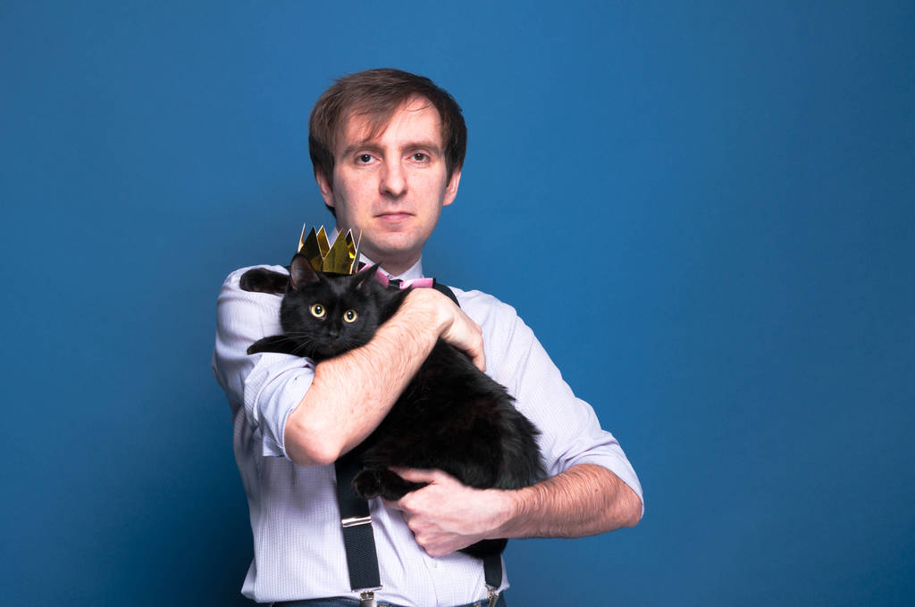 handsome man in shirt and suspender holding cute black cat in golden crown on blue background with copy space - Photo, Image