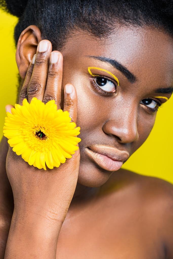 mooie Afro-Amerikaanse meisje met bloem te kijken naar de camera geïsoleerd op geel - Foto, afbeelding