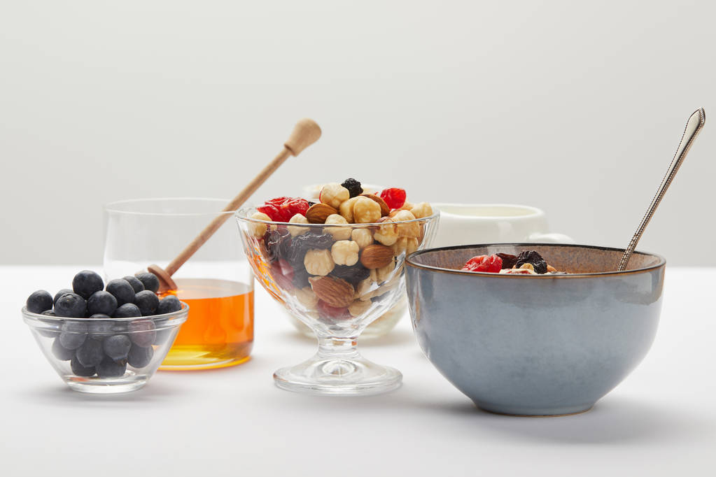 Schalen und Gläser mit Beeren, Nüssen, Honig und Müsli serviert zum Frühstück auf weißem Tisch isoliert auf grau - Foto, Bild