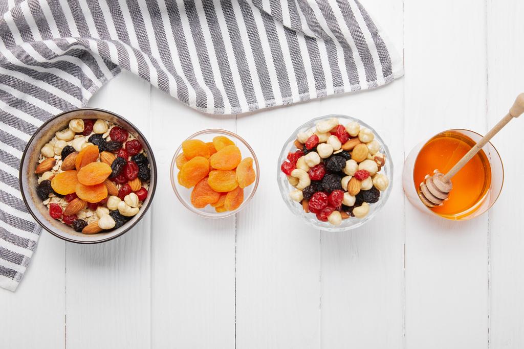 top view of bowls with cereal, dried apricots and berries, honey and nuts on white table with striped cloth - Photo, Image