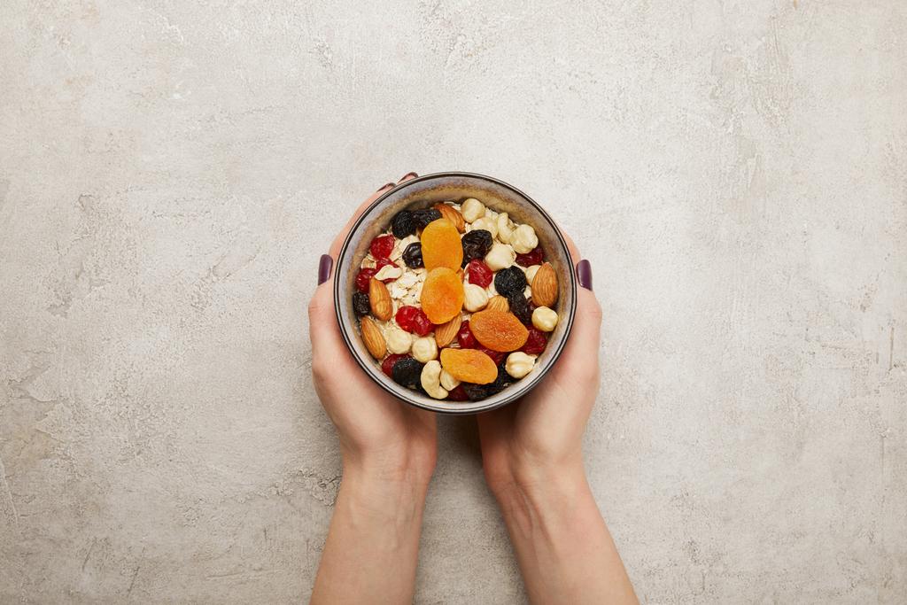 обрізаний вид жінки, що тримає миску з муслі, сушені абрикоси та ягоди, горіхи на текстурованій сірій поверхні
 - Фото, зображення