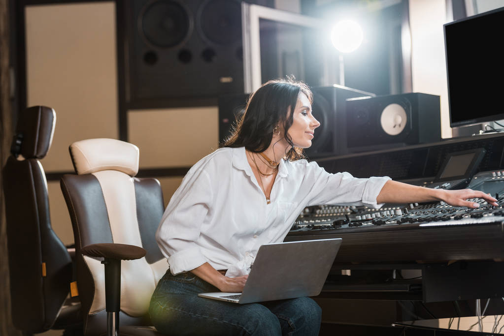atraente, sorrindo produtor de som usando laptop e trabalhando no console de mistura em estúdio de gravação
 - Foto, Imagem