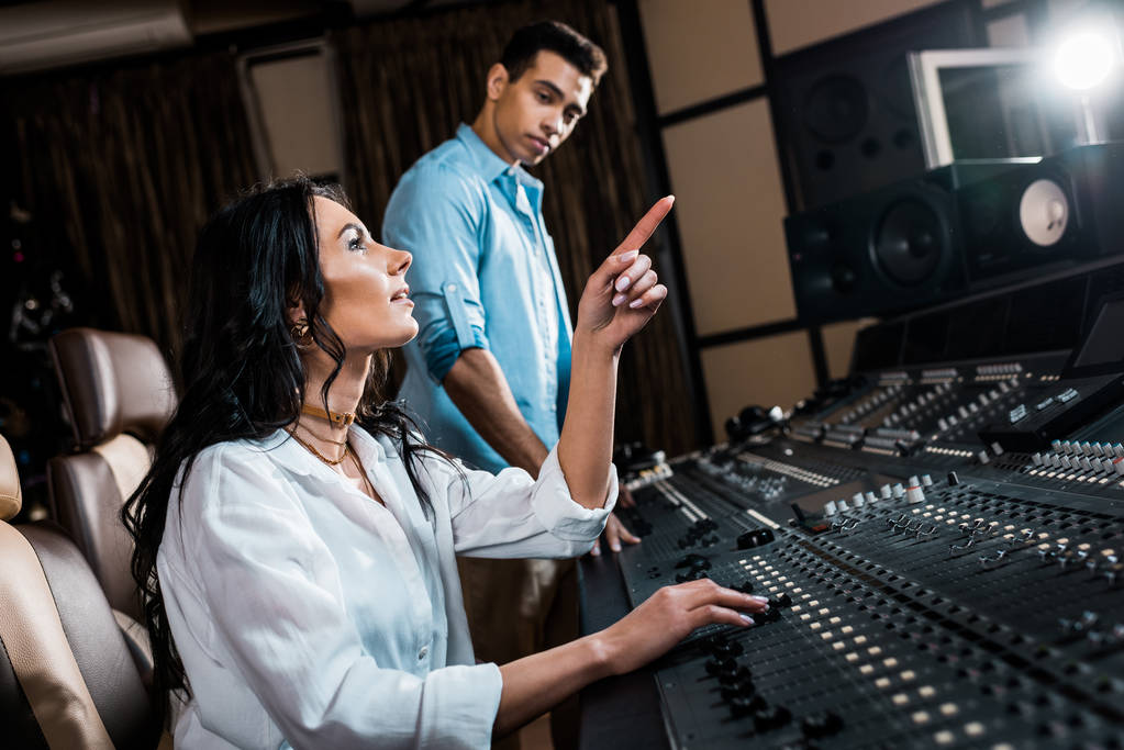hübscher Soundproduzent, der mit dem Finger zeigt, während er im Aufnahmestudio in der Nähe eines Mixed Race Kollegen arbeitet - Foto, Bild
