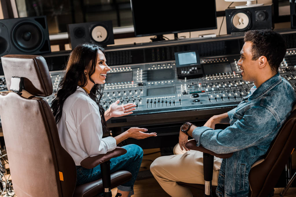 two smiling multicultural sound producers sitting by mixing console in recording studio - Photo, Image