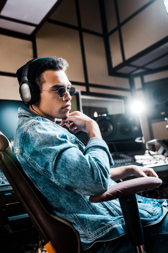 όμορφος σοβαρός παραγωγός μεικτών αγώνων σε γυαλιά ηλίου και ακουστικά που κάθονται στο στούντιο ηχογράφησης - Φωτογραφία, εικόνα