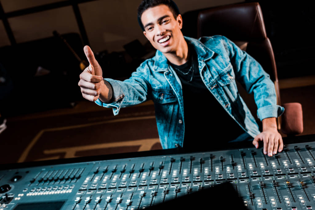 Lächelnder Mixed-Race-Soundproduzent zeigt Daumen hoch, während er neben dem Mischpult im Tonstudio steht - Foto, Bild