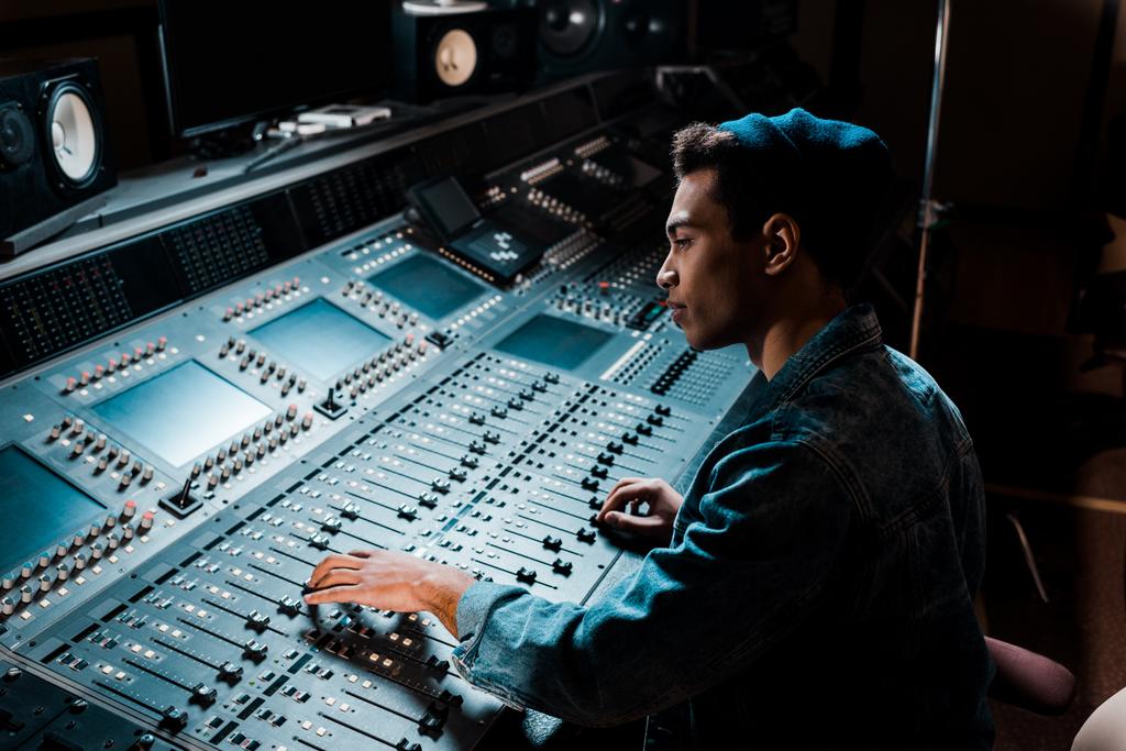 concentrated mixed race sound producer working at mixing console in dark recording studio - Photo, Image
