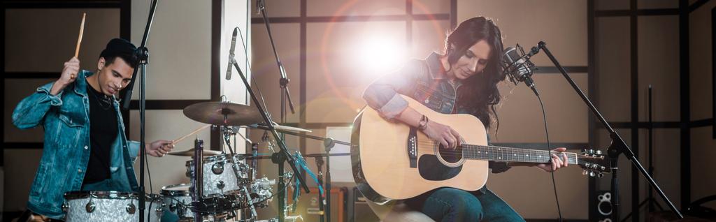 панорамний знімок привабливої жінки, що грає на гітарі, а змішаний расовий музикант грає на барабанах у студії звукозапису
 - Фото, зображення