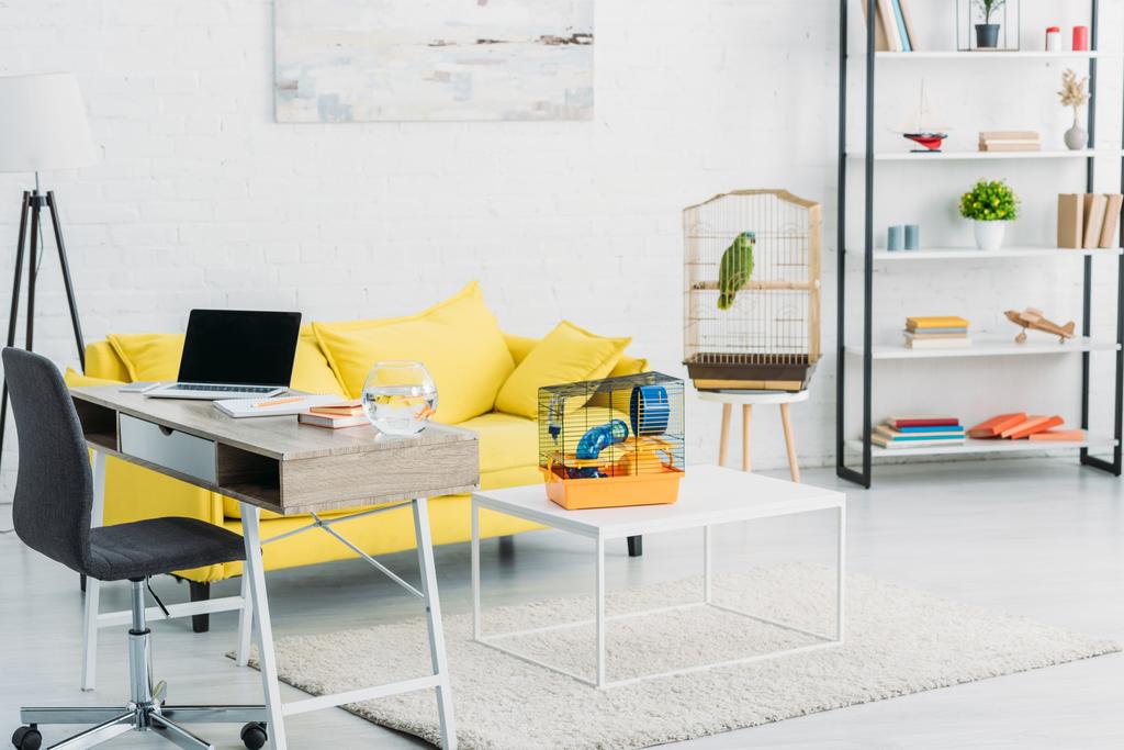 woonkamer met laptop op tafel, gele sofa, groene papegaai in vogelkooi en rek - Foto, afbeelding