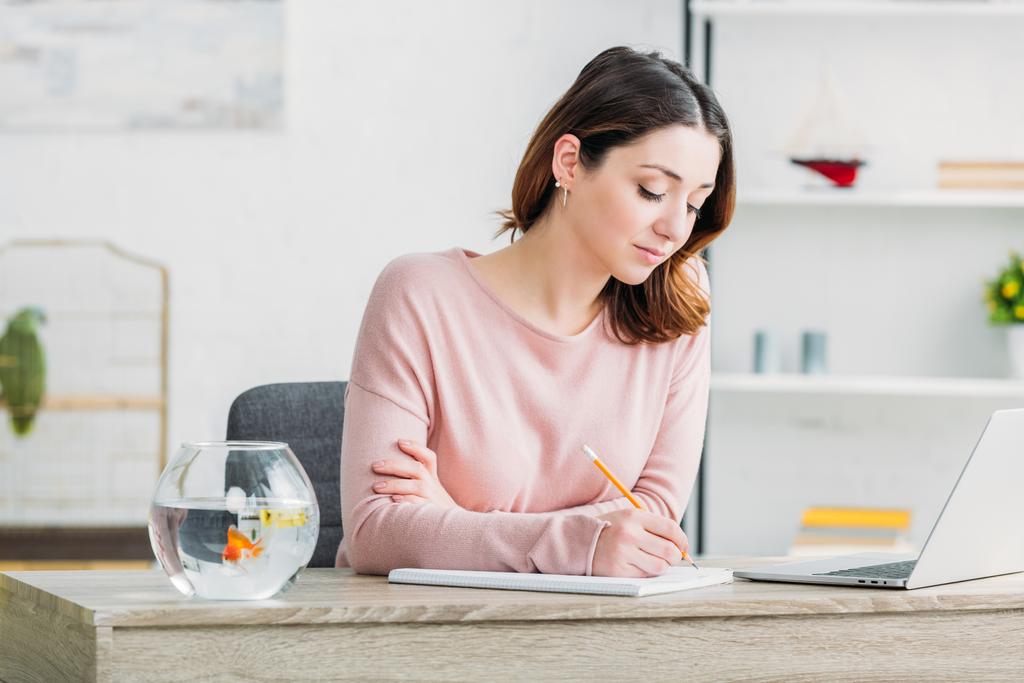 attraktive konzentrierte Frau schreibt in Notizbuch, während sie zu Hause am Tisch sitzt - Foto, Bild