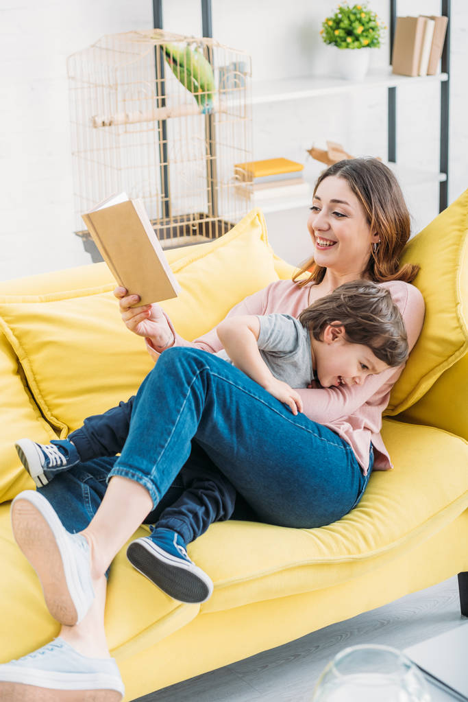 glimlachende moeder met boek plezier met schattige zoon op sofa thuis - Foto, afbeelding
