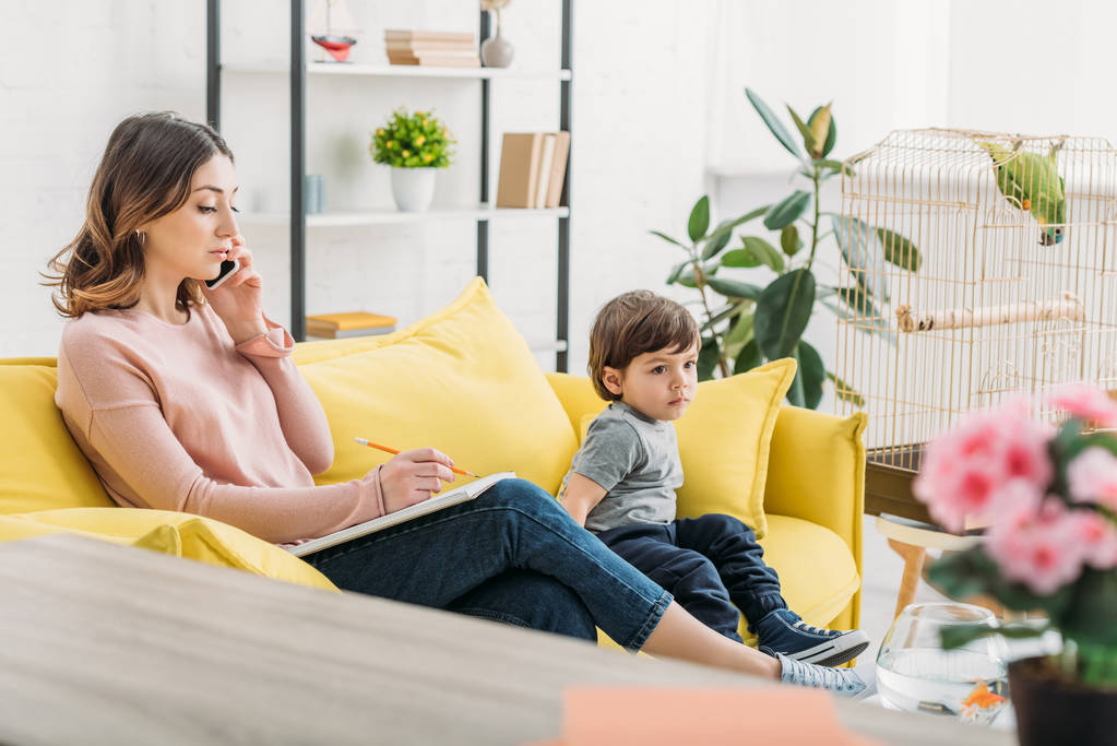 serious woman talking on smartphone while sitting on sofa near adorable son - Photo, Image
