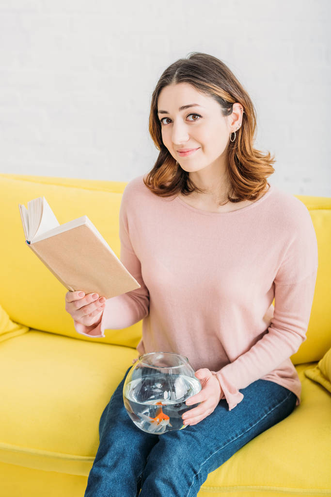 krásná žena, která drží rybí misku a knihu, když sedí na žluté pohovce a dívá se na kameru - Fotografie, Obrázek