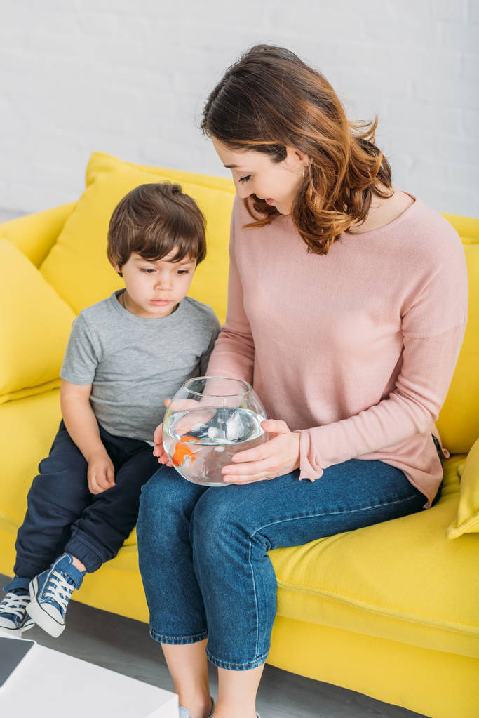 mãe sorridente segurando tigela de peixe enquanto sentado no sofá amarelo com filho adorável
 - Foto, Imagem
