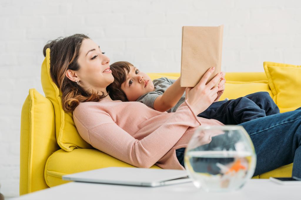 pretty smiling woman  with cute boy reading book while lying on sofa together - Photo, Image
