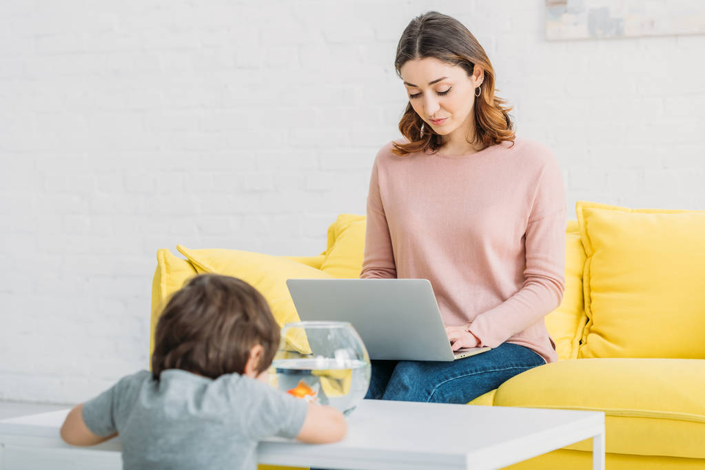 mujer bonita usando el ordenador portátil, mientras que el hijo adorable mirando pecera
 - Foto, imagen