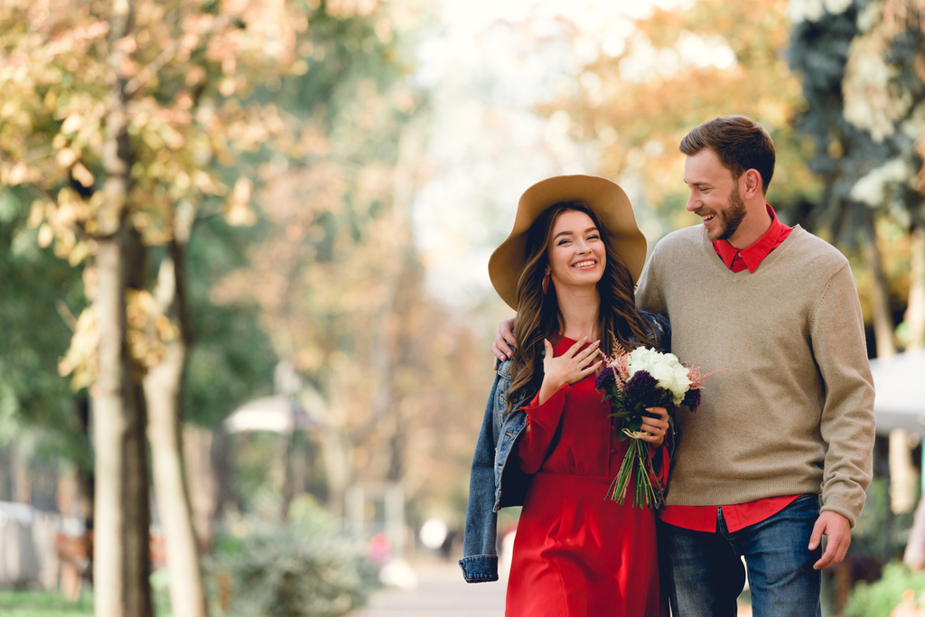 happy man looking at girlfriend in hat smiling while holding flowers  - Photo, Image