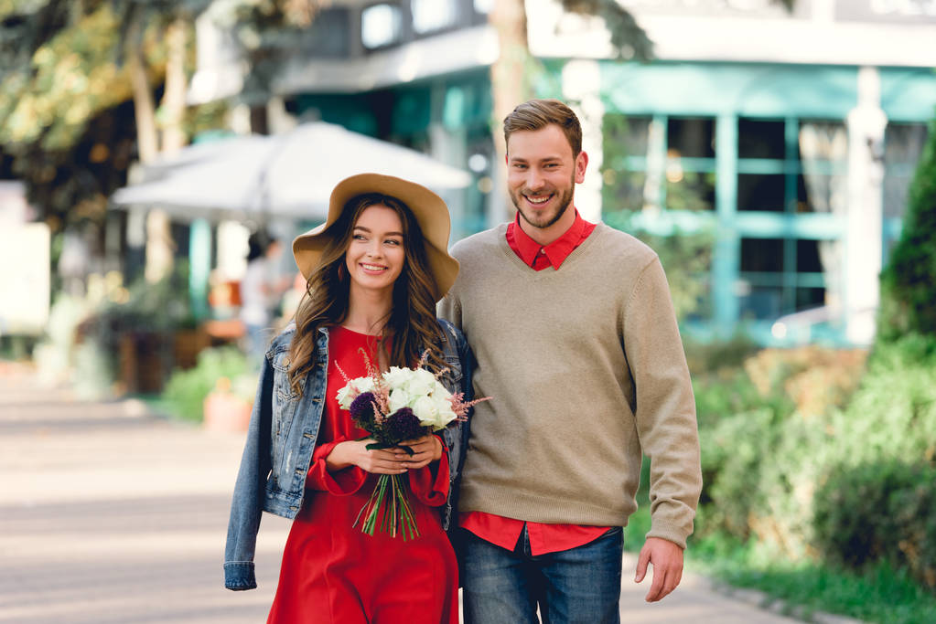 šťastný muž stojící s atraktivní přítelkyní v klobouku, držící květiny  - Fotografie, Obrázek