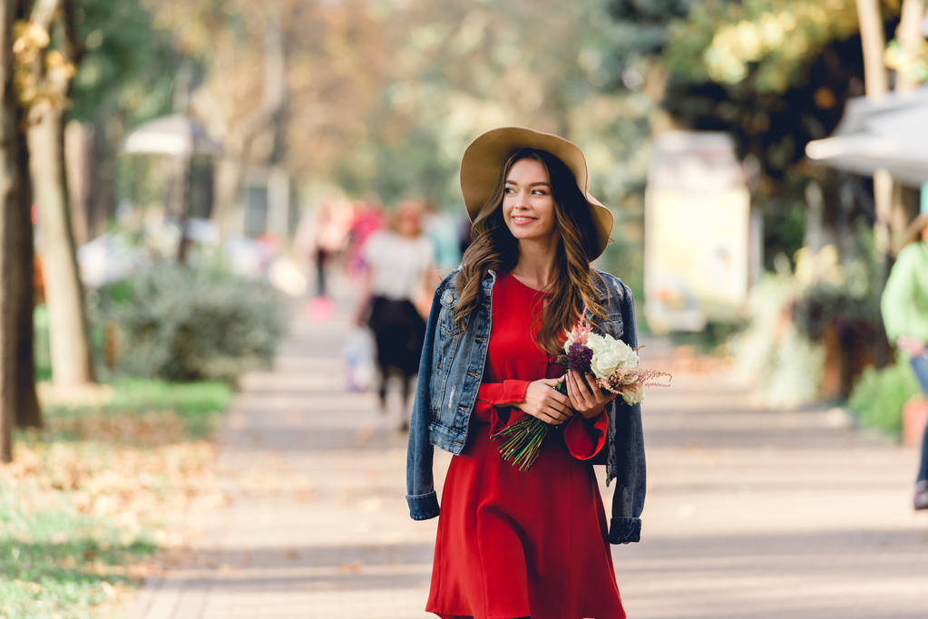 mulher feliz no chapéu sorrindo enquanto segurando flores e caminhando no parque
  - Foto, Imagem