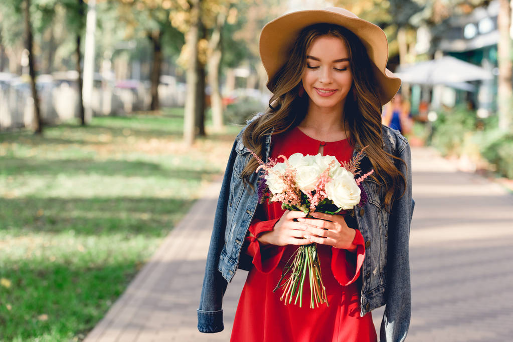 menina atraente no chapéu olhando para as flores, enquanto em pé no parque
  - Foto, Imagem