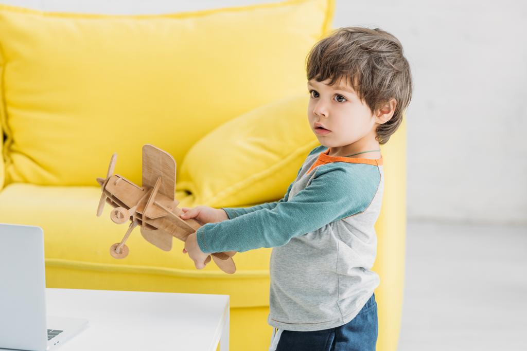 Netter Junge spielt mit hölzernem Flugzeug-Modell in der Nähe von Sofa zu Hause - Foto, Bild