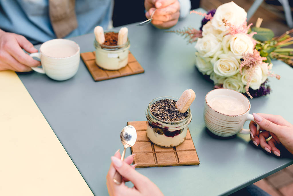 abgeschnittene Ansicht von Mann und Frau mit Löffeln in der Nähe von Desserts und Tassen im Café  - Foto, Bild