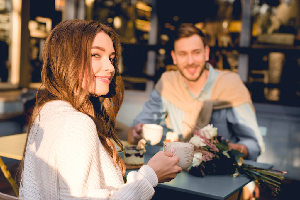 enfoque selectivo de la chica feliz sosteniendo la taza con café mientras está sentado cerca del hombre en la cafetería
  - Foto, imagen