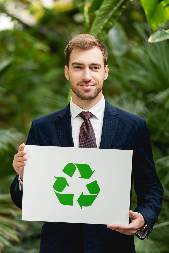 handsome smiling businessman in suit holding card with green recycling sign in greenhouse - Photo, Image