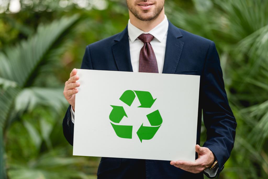 περικομμένη όψη του χαμογελαστή επιχειρηματία στην κάρτα Holding με πράσινο σήμα ανακύκλωσης στο θερμοκήπιο - Φωτογραφία, εικόνα