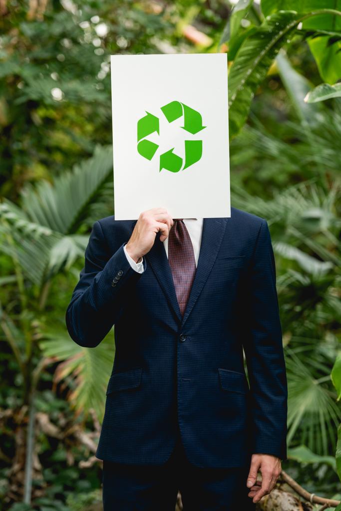 бизнесмен держит карточку с зеленой знак утилизации перед лицом в теплице
 - Фото, изображение