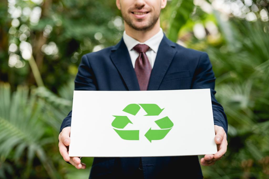 visão parcial do empresário sorridente no cartão de segurança do terno com sinal de reciclagem verde em estufa
 - Foto, Imagem