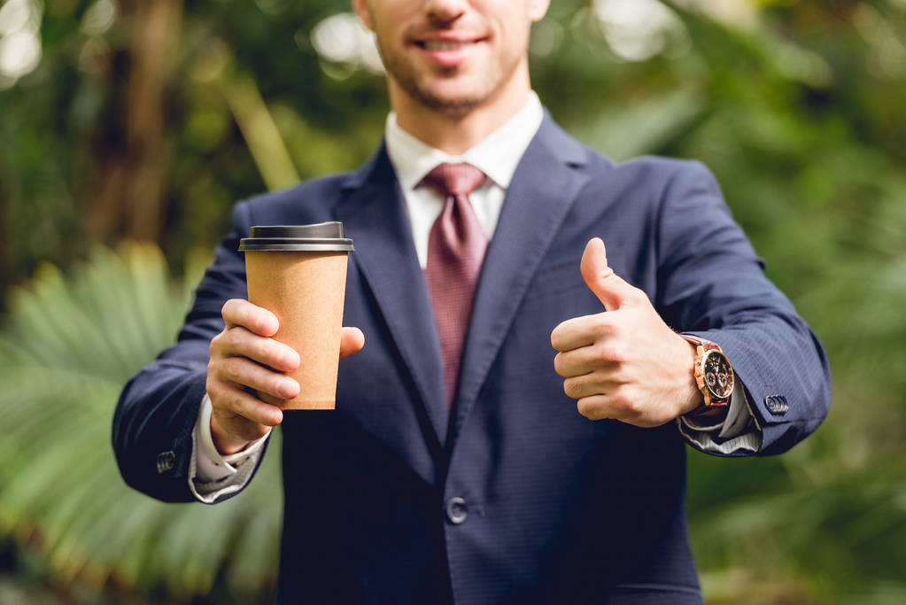bijgesneden uitzicht van lachende zakenman in pak en stropdas houden koffie te gaan en duim omhoog in Oranjerie - Foto, afbeelding