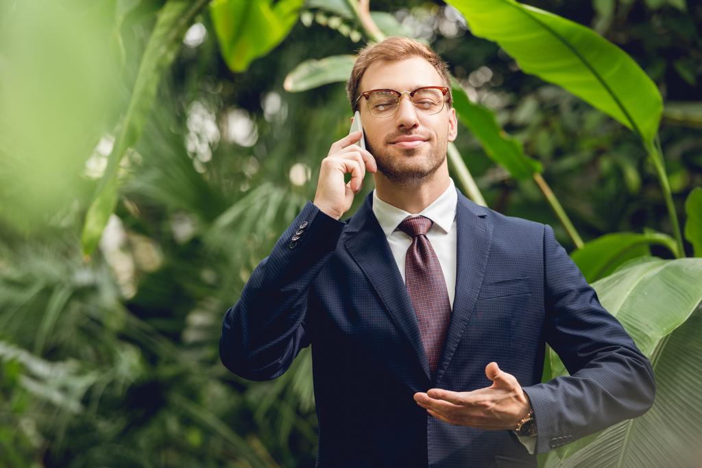 hombre de negocios guapo en traje, corbata y gafas con los ojos cerrados hablando en el teléfono inteligente y respirando aire fresco en invernadero
 - Foto, Imagen
