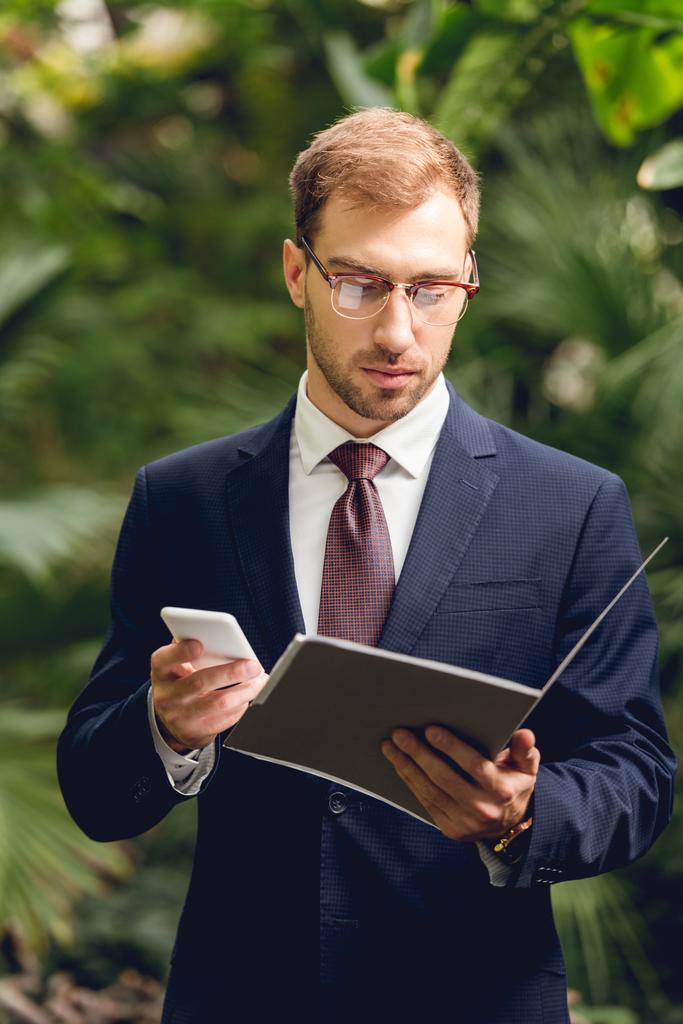 serious businessman in suit using smartphone and holding folder in greenhouse  - Photo, Image