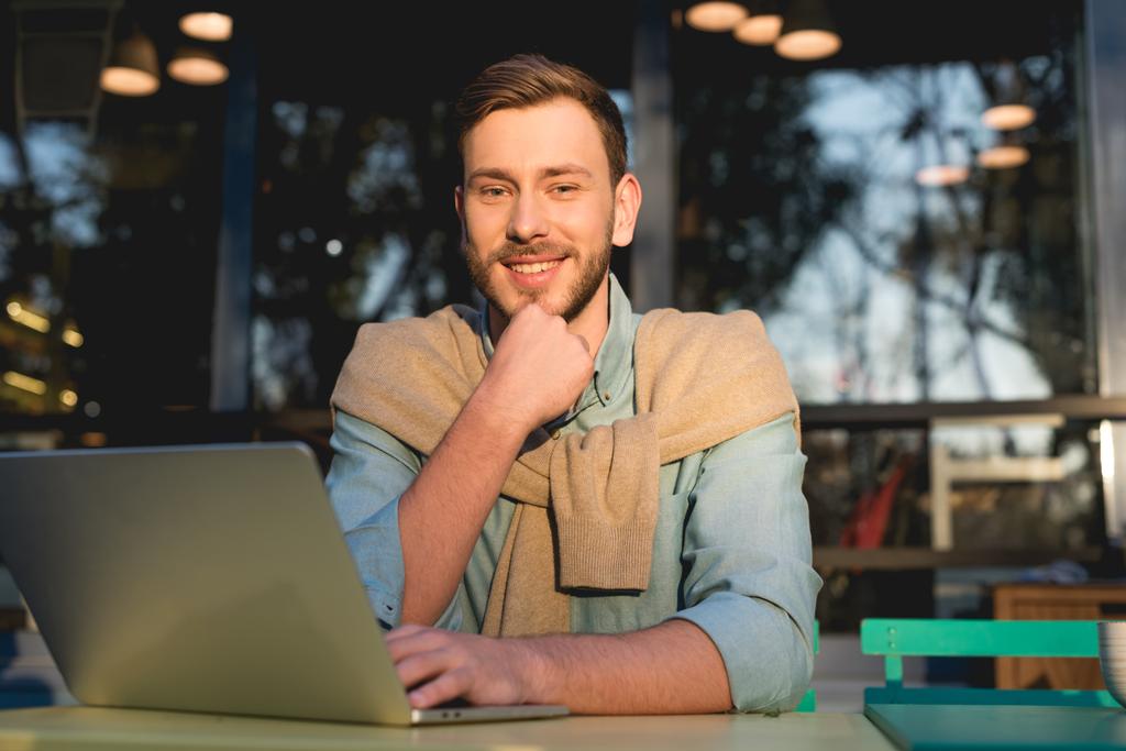 cheerful and handsome freelancer sitting near laptop and looking at camera  - Photo, Image