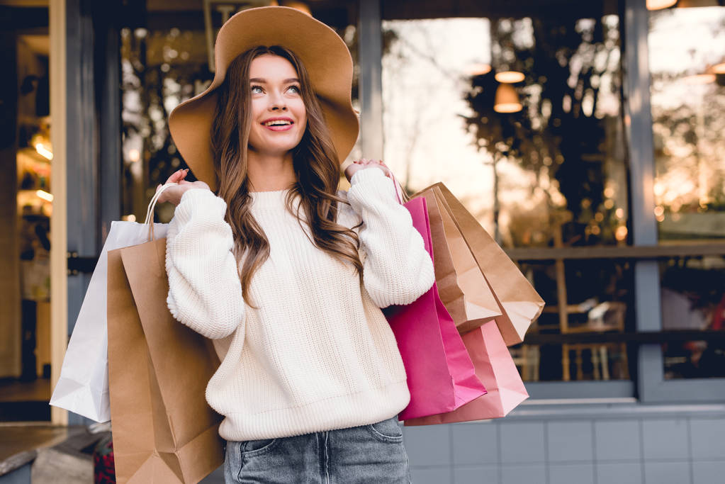glückliche junge Frau mit Hut lächelt, während sie Einkaufstüten in der Hand hält  - Foto, Bild