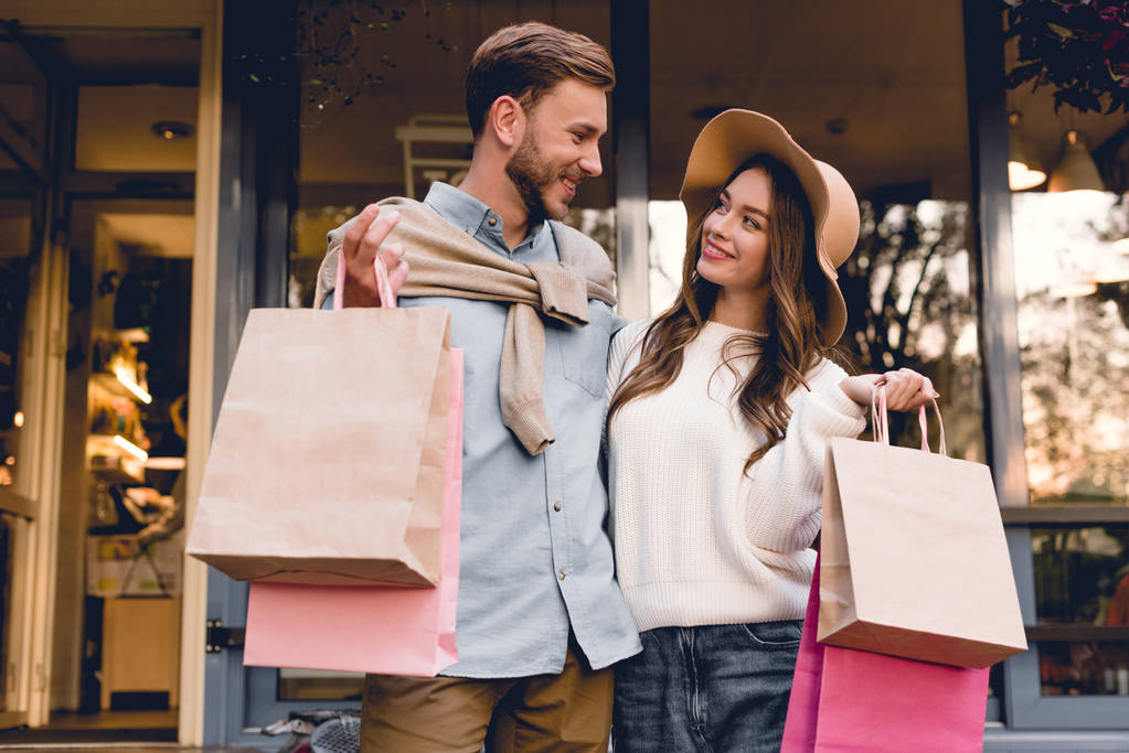vidám szakállas férfi nézi nő kalapban, miközben bevásárló táskák  - Fotó, kép