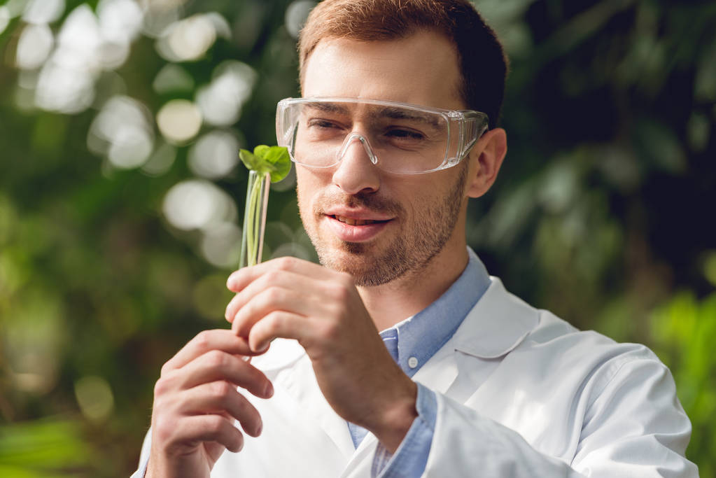 χαμογελαστός όμορφος επιστήμονας με λευκό παλτό και γυαλιά που κρατούν φιάλη με φυτικό δείγμα σε ΠΟΡΤΟΚΑΛΙ - Φωτογραφία, εικόνα