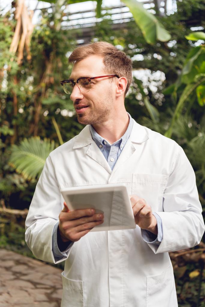 Lächelnder Wissenschaftler in weißem Mantel und Brille mit digitalem Tablet in der Orangerie - Foto, Bild