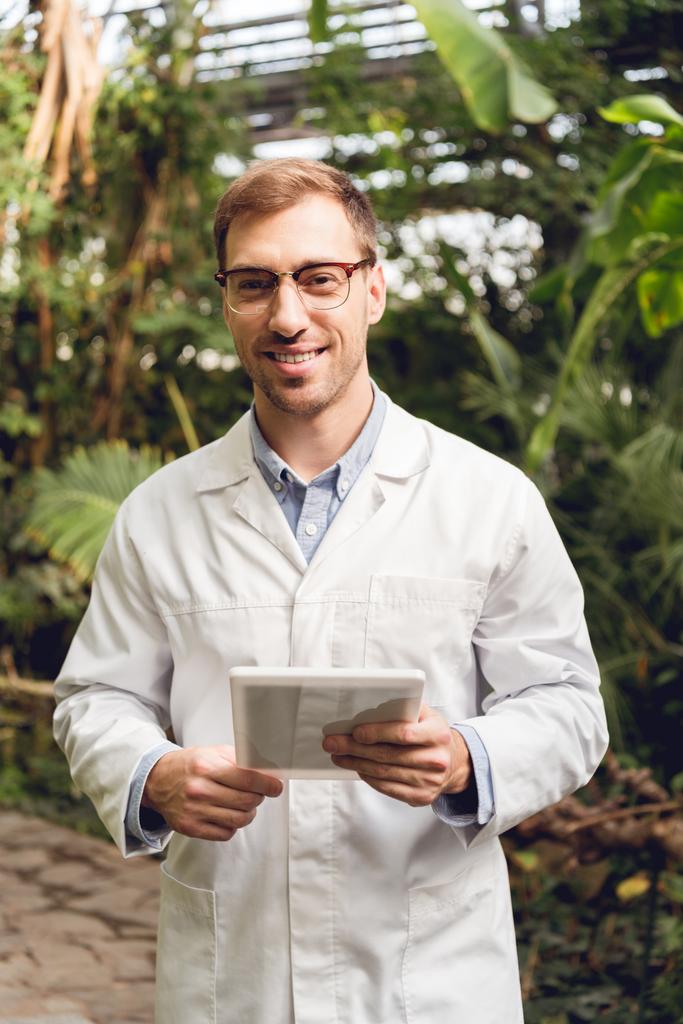 Улыбающийся красивый ученый в белом халате и очках держит цифровую табличку в оранжерее
 - Фото, изображение